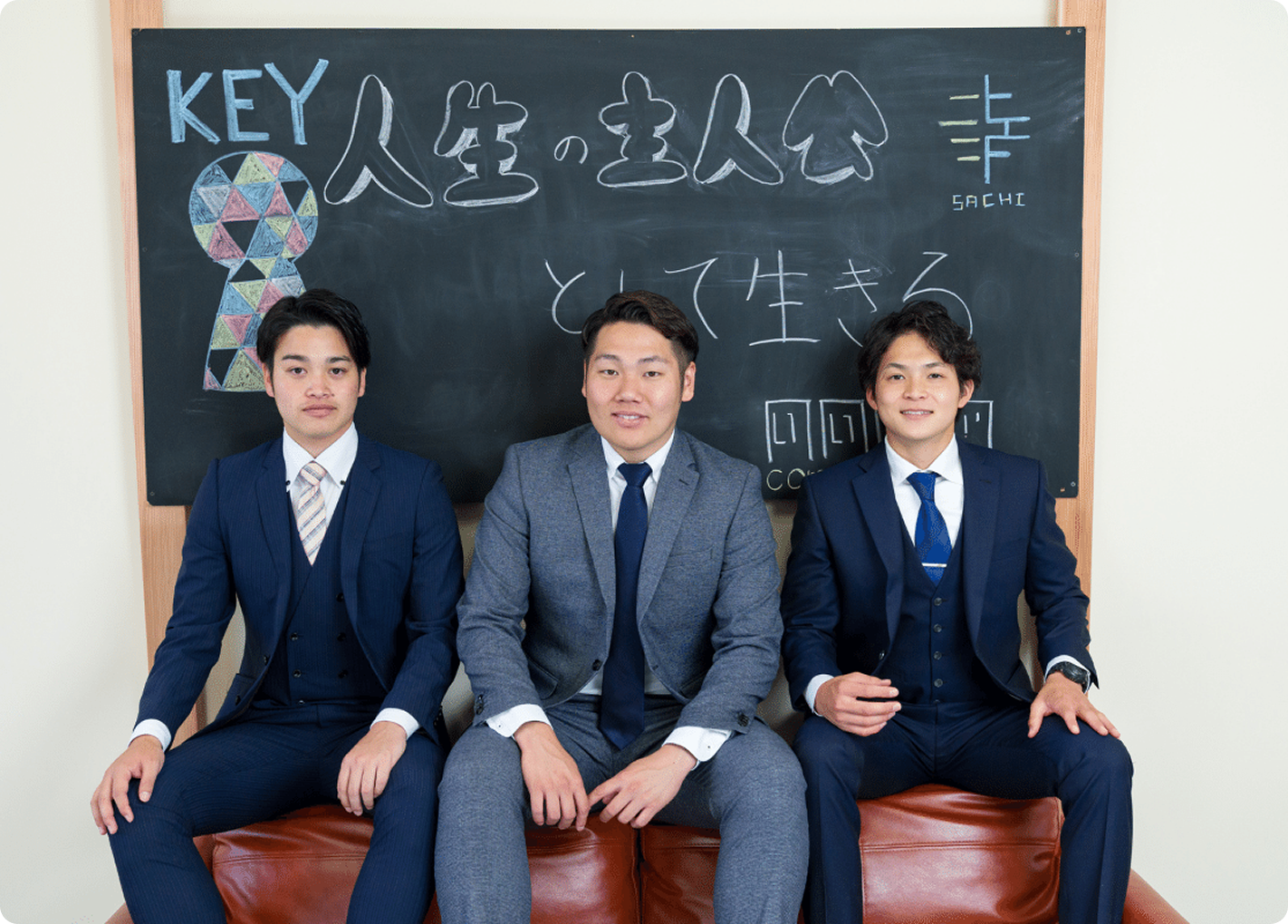 スーツ姿の男性3名が黒板の前に座っている写真。黒板には『人生の主人公として生きる』と書かれている