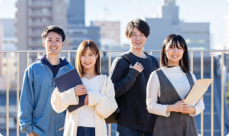 学生らしい若者4名が笑顔で並んでいる様子。屋外の明るい雰囲気